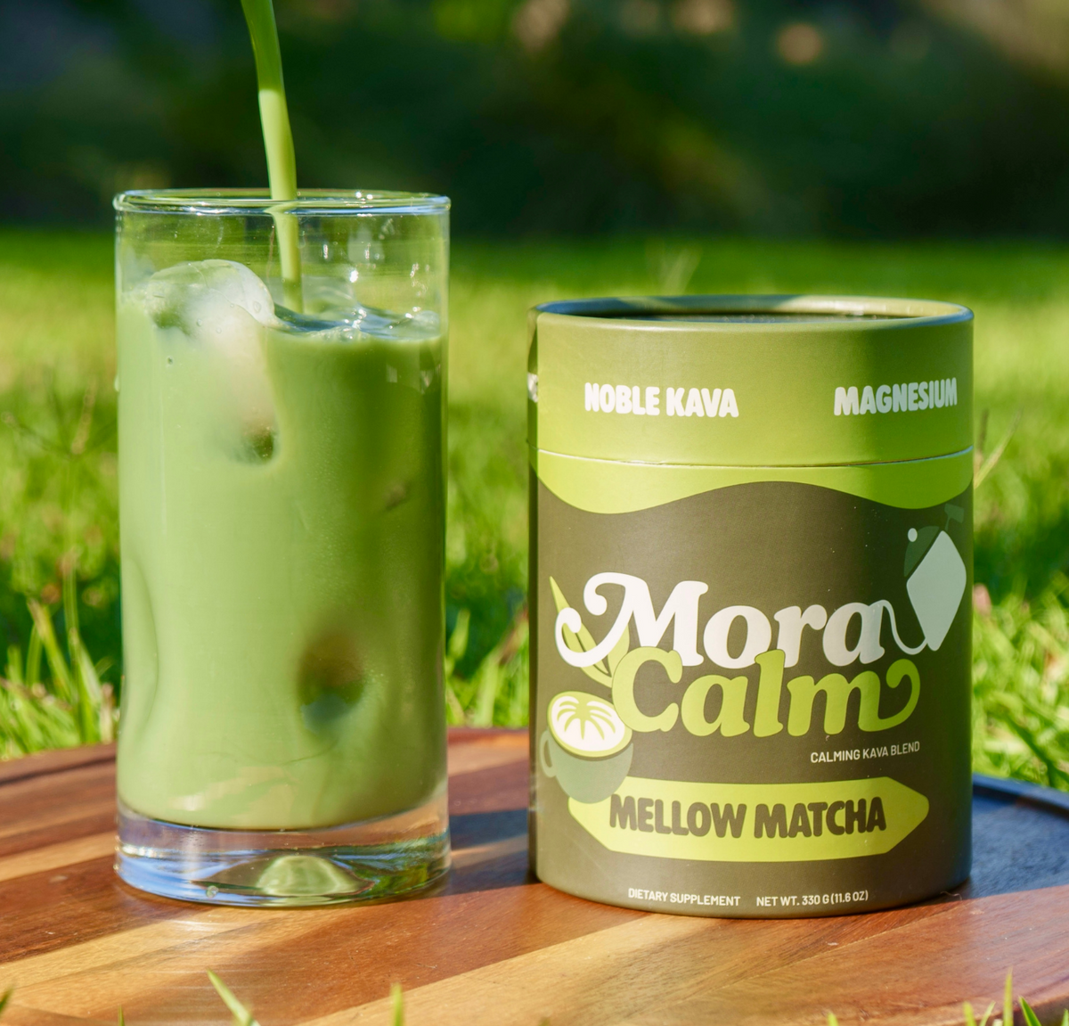 A glass of iced Mora Calm matcha sits beside a cylindrical canister of Mora Calm Mellow Matcha on a wooden tray outdoors on grass.